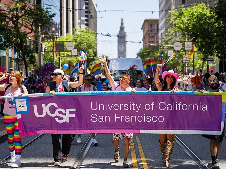 With the Ferry Terminal clock tower seen in the distance, UCSF community members participate as a contingent in the 2025 San Francisco Pride parade, progressing down Market Street behind a UCSF banner held aloft by several colorfully clothed members.